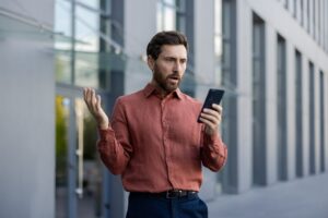 Mature businessman outdoors looking surprised at smartphone in urban setting, wearing smart casual